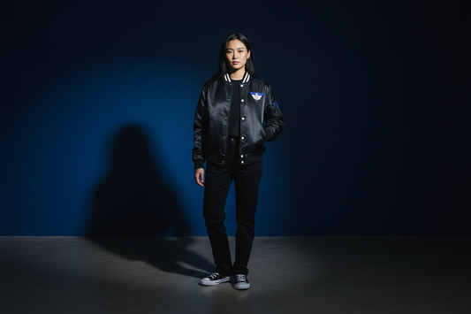 A woman stands in a focused spotlight wearing a black satin varsity jacket with a lion emblem. The deep blue background and dramatic lighting create a mood of quiet luxury, confidence, and silent authority—core to the R10 identity.