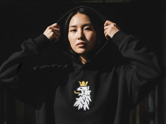 Model wearing a black R10 lion-crest hoodie in a dark industrial warehouse. Minimalist streetwear style with premium embroidery and quiet-luxury aesthetic.