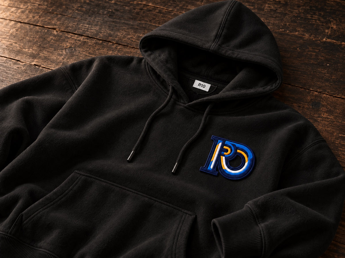 R10 Monogram hoodie on dark timber, showing its hood seam, white neck label, drawcord, embroidery, pocket, and ribbing.