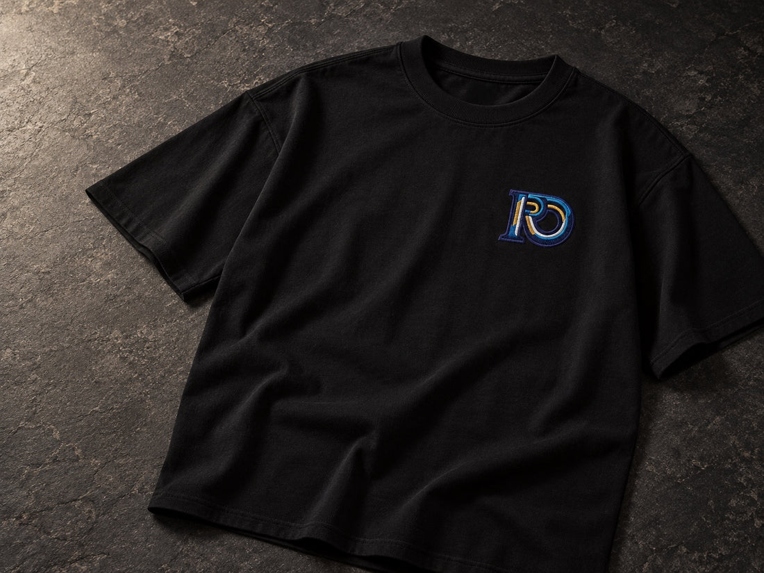 Black R10 Monogram Tee laid on dark stone, showing the collar, sleeves, and chest embroidery.