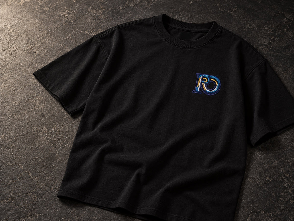Black R10 Monogram Tee laid on dark stone, showing the collar, sleeves, and chest embroidery.